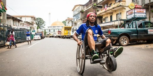 A man travels along a street in his wheelchair during a three-day lockdown to prevent the spread on the Ebola virus, Freetown, Sierra Leone, Sept. 21, 2014 (AP photo by Michael Duff).