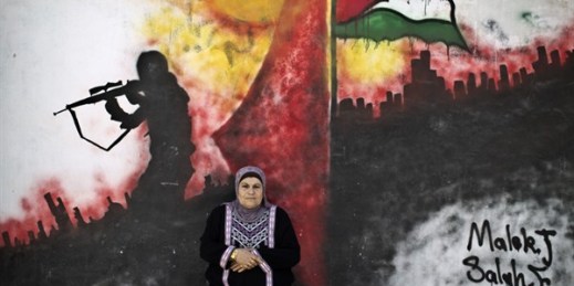 A Palestinian refugee poses for a picture in front of a wall painted with a mural in the Kalandia refugee camp between Jerusalem and the West Bank city of Ramallah, June 18, 2014 (AP photo by Muhammed Muheisen).