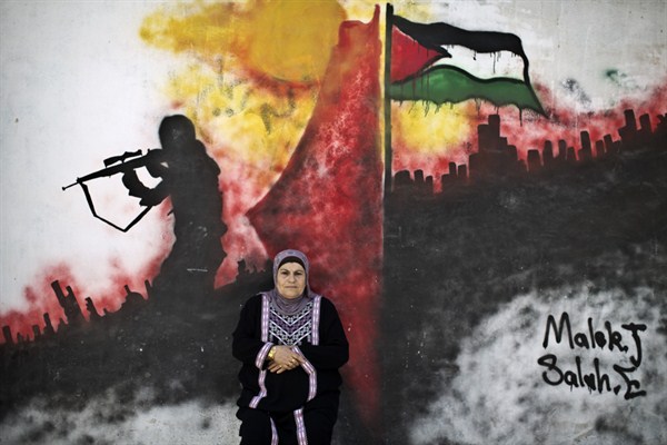 A Palestinian refugee poses for a picture in front of a wall painted with a mural in the Kalandia refugee camp between Jerusalem and the West Bank city of Ramallah, June 18, 2014 (AP photo by Muhammed Muheisen).