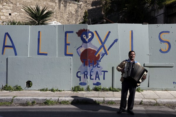 A busker plays music in front of graffiti that refers to Greece’s Prime Minister Alexis Tsipras in the Plaka district of Athens, Feb. 21, 2015 (AP photo by Thanassis Stavrakis).