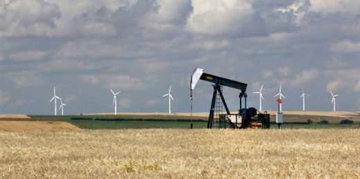 An oil field pump works while wind turbines generate power in the distance, Saskatchewan, Canada, Aug. 29, 2014 (AP photo by Larry MacDougal).