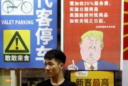 A man sits in front of a restaurant billboard stating that American customers will be charged 25 percent extra due to ongoing China-U.S. trade tensions, Guangzhou, China, Aug. 13, 2018 (Chinatopix photo via AP Images).