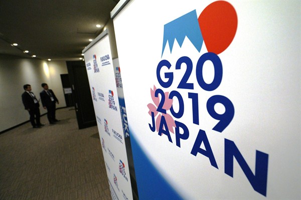 Staff members stand near the emblem for the 2019 Group of 20 leaders’ summit at the entrance of the press center of the G-20 Finance Ministers’ and Central Bank Governors’ meeting, in Fukuoka, Japan, June 9, 2019 (AP photo by Eugene Hoshiko).