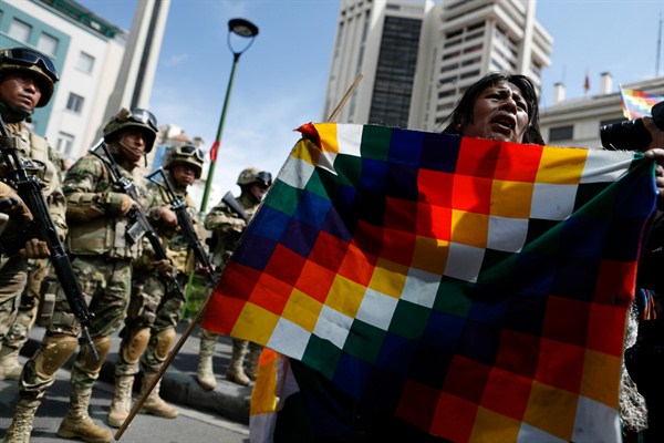 A supporter of former President Evo Morales holds a Wiphala flag, an emblem of the indigenous people of the Andes region, in front of soldiers blocking a street in downtown La Paz, Bolivia, Nov. 15, 2019 (AP photo by Natacha Pisarenko).