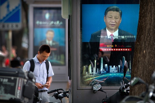 A man looks at his smartphone near video display screens showing Chinese President Xi Jinping, in Beijing, Aug. 22, 2018 (AP photo by Mark Schiefelbein).