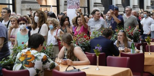 People protesting against Italy’s COVID-19 Green Pass walk past customers sitting at a cafe in the Galleria Vittorio Emanuele shopping arcade, Milan, Italy, July 24, 2021 (AP photo by Antonio Calanni).