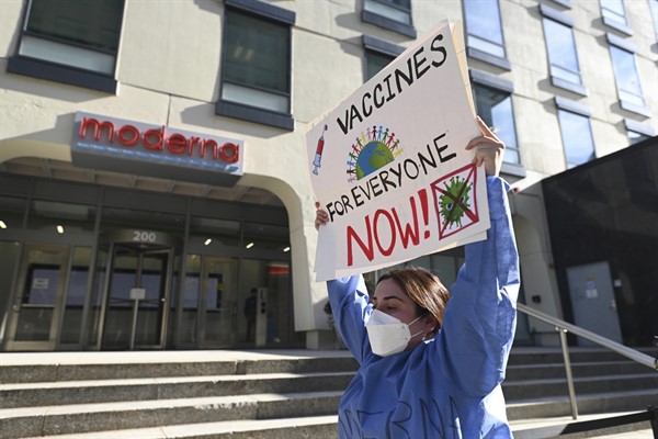 A protester supporting the “Vaccinate Our World” campaign holds a sign in front of the world headquarters of COVID-19 vaccine maker Moderna, Cambridge, Mass, Nov. 18, 2021 (photo by Gretchen Ertl for AIDS Healthcare Foundation via AP Images).