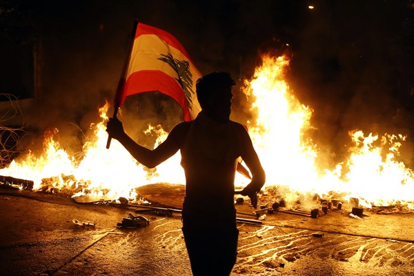 An anti-government protester waves a Lebanese flag in front of a burning barricade on a road leading to the parliament building, Beirut, Lebanon, Nov. 13, 2019 (AP photo by Bilal Hussein).