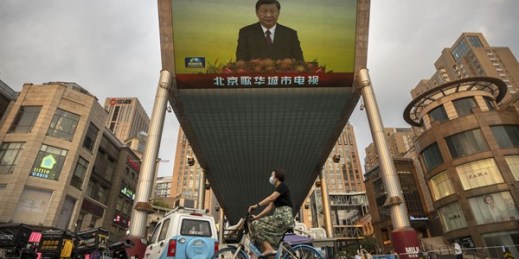 A woman wearing a face mask rides a bicycle past a large television screen displaying Chinese President Xi Jinping, Hong Kong, July 1, 2022 (AP photo by Mark Schiefelbein).