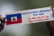 A protester holds a sign that reads in French, “Restitution and Reparations for Haiti”.