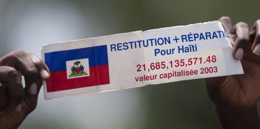 A protester holds a sign that reads in French, “Restitution and Reparations for Haiti”.