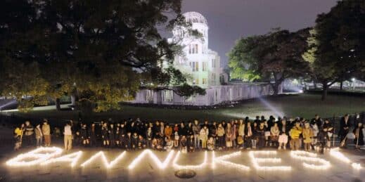 A group of people stand behind lit candles arranged to read “Ban Nukes!”.
